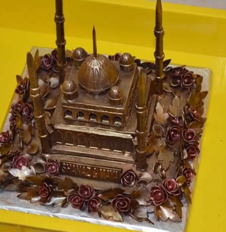 A square chocolate cake built as a mosque, with a ribbed dome, four tall minarets and a ring of dark red chocolate roses, the couple’s names engraved on a plaque at the front