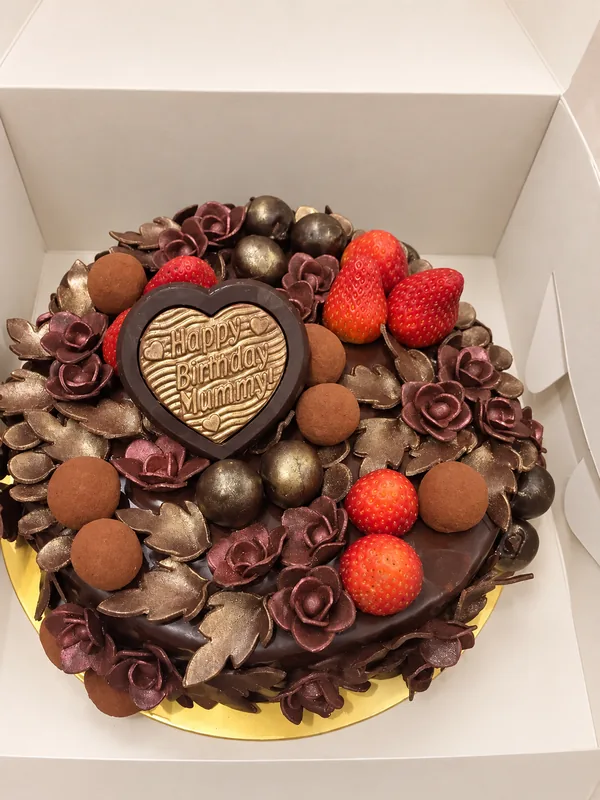 A chocolate-covered birthday cake topped with chocolate roses, bronze leaves, truffles and fresh strawberries, with a chocolate heart plaque reading Happy Birthday Mummy